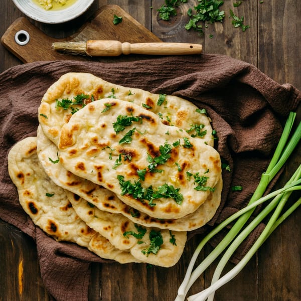 Stack of fresh Indian naan bread topped with cilantro, alongside green onions and a brush.